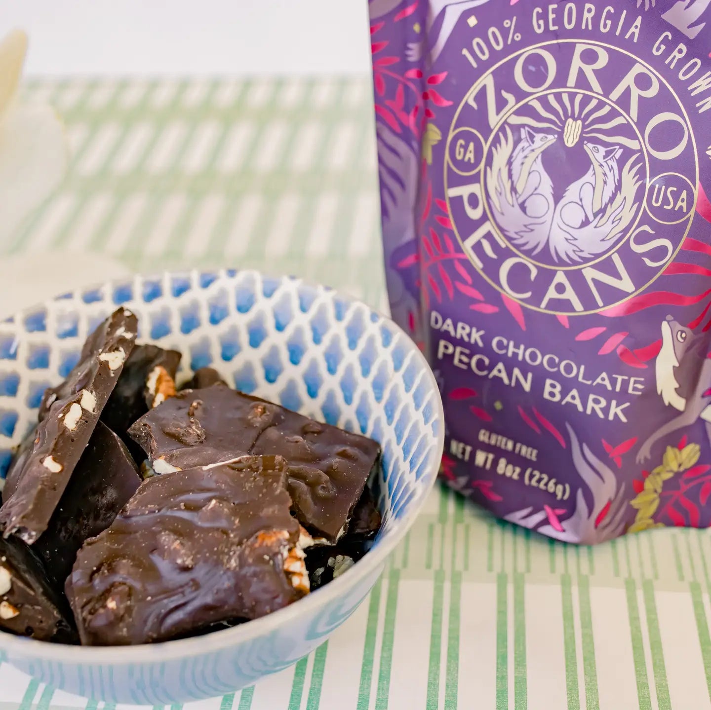 Zorro Pecans dark chocolate pecan bark pieces in a blue bowl beside purple bag
