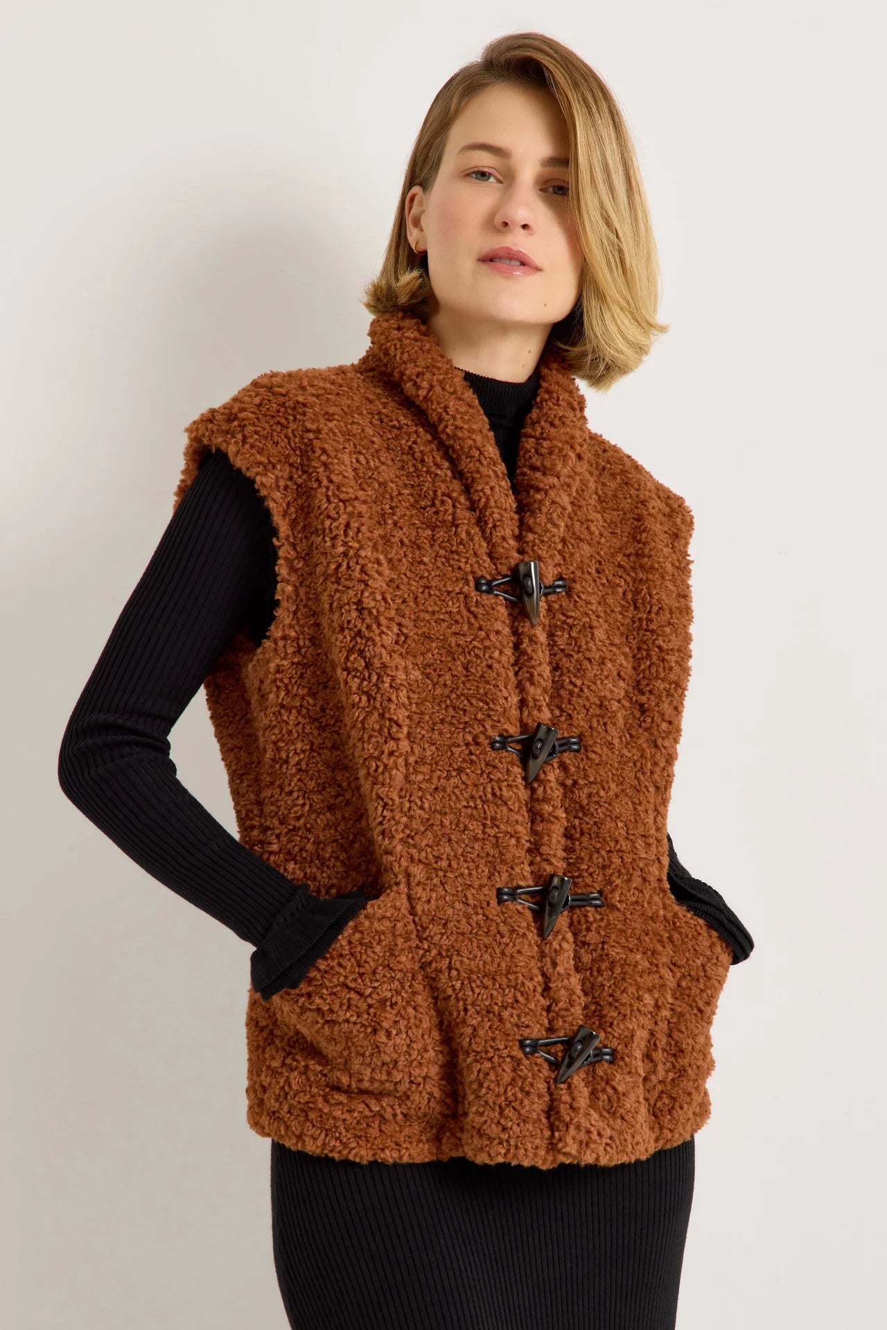 Brown sherpa fleece vest with black toggle closures worn over black ribbed knit