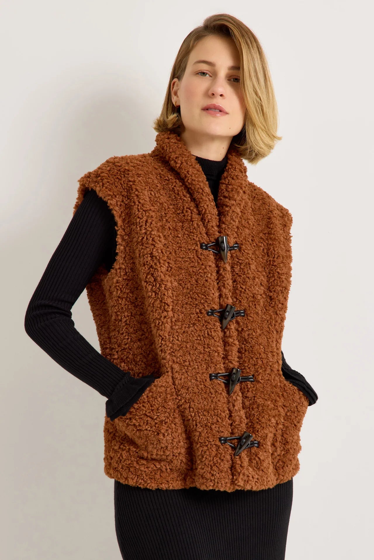 Brown sherpa fleece vest with black toggle closures worn over black ribbed knit